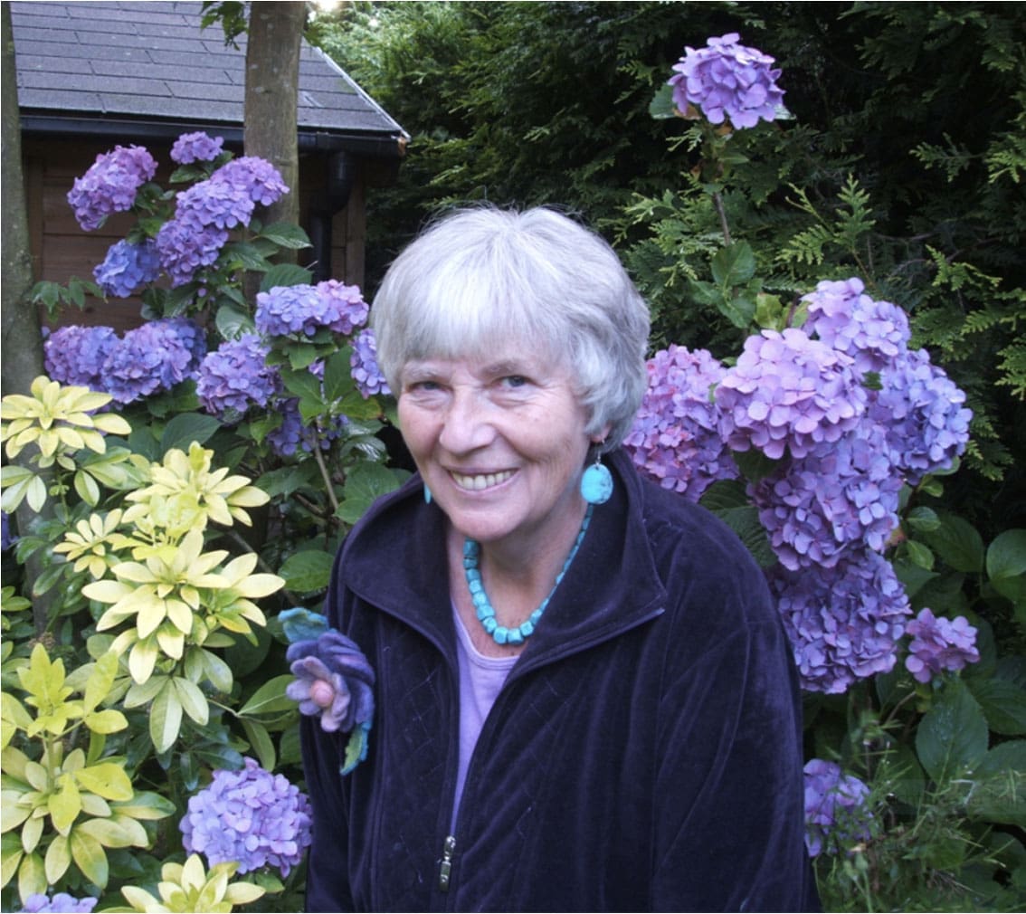 Annelies Spaargaren lachend in een tuin vol paarse en gele bloemen, symbool voor groei en transformatie.